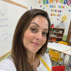 Maria Nidia sorrindo numa sala de aula infantil, com mesas e cadeiras coloridas ao fundo