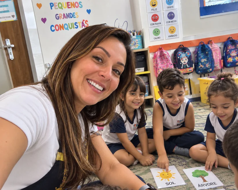 Maria Nidia sorrindo, de avental, sentada no chão da sala com três crianças de uniforme em volta de cartões ilustrados